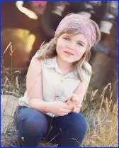 A little girl sitting in the grass wearing jeans and a headband.